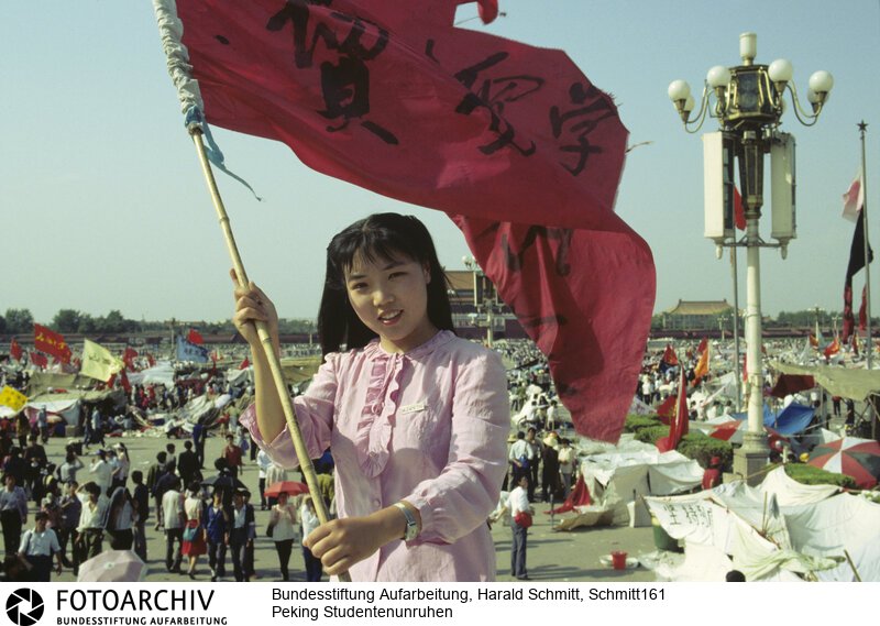 Das Gesicht der Revolte. Furchtlos und zuversichtlich schwenkt die 20jährige Biologiestudentin Xia die Fahne ihrer Hochschule über dem "Platz des Himmlischen Friedens"; auf dem die Demonstranten ihr Lager aufgeschlagen haben. Sie kämpft nicht gegen den Sozialismus; aber gegen Unfreiheit. Dia: 56/05 <BR>Aufnahmedatum: Mai / Juni 1989<BR>Material/Technik: Foto / Dia<BR>Aufnahmeort: Peking / China<BR>Signatur: Schmitt161