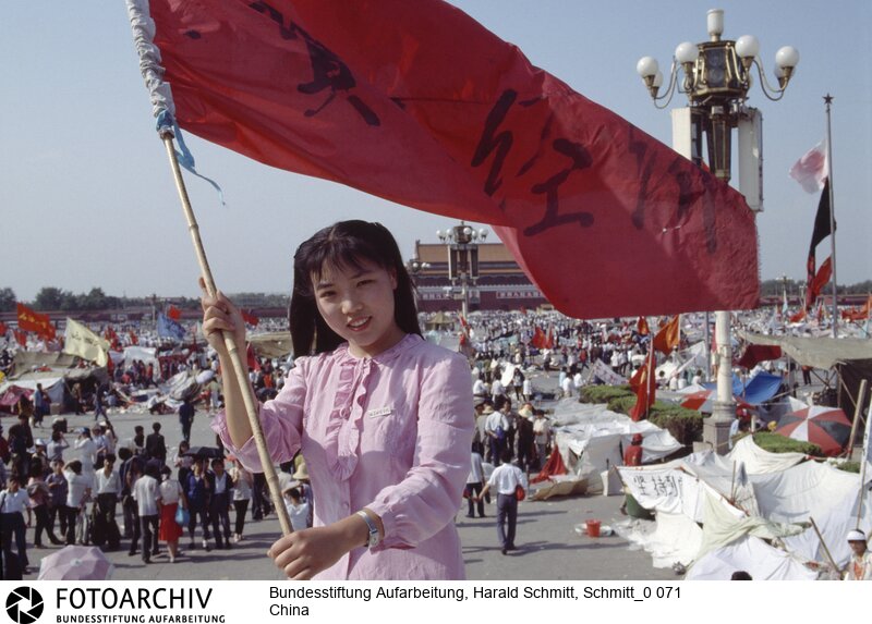 Das Gesicht der Revolte. Furchtlos und zuversichtlich schwenkt die 20jährige Biologiestudentin Xia die Fahne ihrer Hochschule über dem "Platz des Himmlischen Friedens", auf dem die Demonstranten ihr Lager aufgeschlagen haben. Sie kämpft nicht gegen den Sozialismus, aber gegen Unfreiheit<BR>Aufnahmedatum: 1989<BR>Material/Technik: Foto<BR>Aufnahmeort: Peking / China<BR>Signatur: Schmitt_0 071