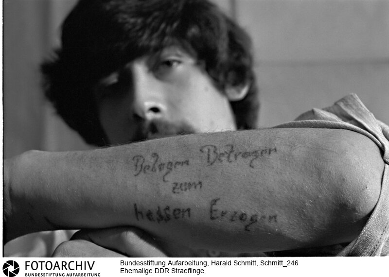 Tätowierung auf der Brust von Peter WIESNER, eines ehemaligen DDR Sträflings, der wegen seines Fluchtversuches einsitzen musste. Mit Tinte und einer Nadel wurde: Belogen, Betrogen zum Hassen erzogen auf den Arm tätowiert. Das machen angeblich sehr viele Häftlinge. Film: 81 0119  Negativ: 33<BR>Aufnahmedatum: 1981<BR>Material/Technik: Foto<BR>Aufnahmeort: DDR (Deutschland)<BR>Signatur: Schmitt_246