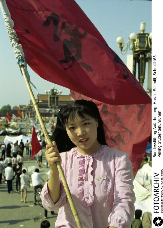 Das Gesicht der Revolte. Furchtlos und zuversichtlich schwenkt die 20jährige Biologiestudentin Xia die Fahne ihrer Hochschule über dem "Platz des Himmlischen Friedens", auf dem die Demonstranten ihr Lager aufgeschlagen haben. Sie kämpft nicht gegen den Sozialismus; aber gegen Unfreiheit. Dia: 56/05 <BR>Aufnahmedatum: Mai / Juni 1989<BR>Material/Technik: Foto / Dia<BR>Aufnahmeort: Peking / China<BR>Signatur: Schmitt163