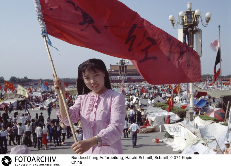 Das Gesicht der Revolte. Furchtlos und zuversichtlich schwenkt die 20jährige Biologiestudentin Xia die Fahne ihrer Hochschule über dem "Platz des Himmlischen Friedens"; auf dem die Demonstranten ihr Lager aufgeschlagen haben. Sie kämpft nicht gegen den Sozialismus; aber gegen Unfreiheit<BR>Aufnahmedatum: 1989<BR>Material/Technik: Foto<BR>Aufnahmeort: Peking / China<BR>Signatur: Schmitt_0 071 Kopie