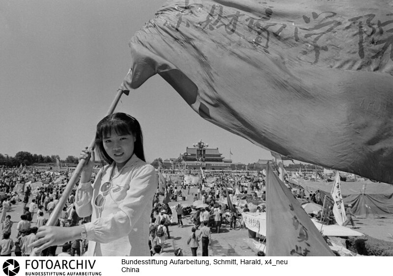 Das Gesicht der Revolte. Furchtlos und zuversichtlich schwenkt die 20jährige Biologiestudentin Xia die Fahne ihrer Hochschule über dem "Platz des Himmlischen Friedens", auf dem die Demonstranten ihr Lager aufgeschlagen haben. Sie kämpft nicht gegen den Sozialismus, aber gegen Unfreiheit.<BR>Aufnahmedatum: Mai / Juni 1989<BR>Material/Technik: Foto<BR>Aufnahmeort: Peking / China<BR>Signatur: x4_neu