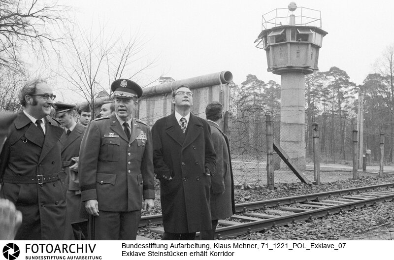 Foto (v.l.): Bezirksbürgermeister Wolfgang Rothkegel (CDU), General William Cobb, Kommandeur US Brigade, Regierender Bürgermeister Klaus Schütz (SPD) an der durch die Exklave führenden Reichsbahntrasse. Berlin (Berlin West), 21. 12. 1971. Nach dem gestern geschlossenem Abkommen besuchten Politiker und US-Militär per Hubschrauber die Einwohner der Exklave. Die DDR wird einen 20 Meter breiten Geländestreifen auf gut einem Kilometer abtreten. Der Senat baut darauf eine Straße, die das Eremitendasein der Bewohner beendet.<BR>Material/Technik: Foto<BR>Berlin (West)<BR>Signatur: 71_1221_POL_Exklave_07