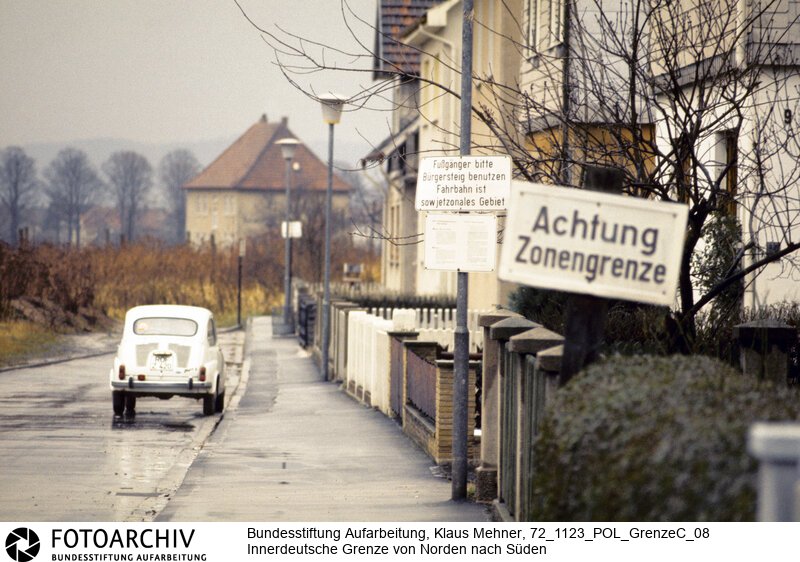 Landkreis Hersfeld-Rothenburg (Hessen), 23. 11. 1972. Seit dem Bau der Mauer hat die Deutsche Demokratische Republik (DDR) die Demarkationslinie entlang ihrer Staatsgrenze West optimiert. Die dem Gelände angepasste Zonengrenze zementiert die Teilung Deutschlands, für Flüchtlinge besteht Lebensgefahr. Das durch Grenzsoldaten bewachte Sperrsystem ist durch Minen, Todesstreifen und Wachtürme abgeriegelt. Foto: Im Hessischen Obersuhl kann schadlos in der Zone geparkt werden. In the village of Obersuhl belongs the road to East Germany.<BR>Material/Technik: Foto<BR>Hessen<BR>Signatur: 72_1123_POL_GrenzeC_08