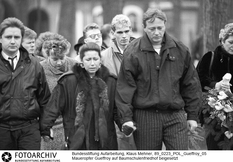 Berlin (Bezirk Berlin) DDR, 23. 02. 1989. Das letzte Opfer des Schießbefehls an der Staatsgrenze der Deutschen Demokratischen Republik (DDR), Chris Gueffroy starb in der Nacht zum 06. Februar 1989 durch Schüsse von Grenzposten bei einem Fluchtversuch an der Berliner Mauer. Die Beisetzung fand auf dem Baumschulenweg-Friedhof statt. Ein Grossaufgebot von Kräften des Ministeriums für Staatssicherheit (MfS, Stasi) und der Volkspolizei (VP, VoPo) schirmte die Trauernden ab. Foto: Trauergemeinde, in der Mitte die Mutter Karin Gueffroy.<BR>Material/Technik: Foto<BR>Bezirk Berlin<BR>Signatur: 89_0223_POL_Gueffroy_05