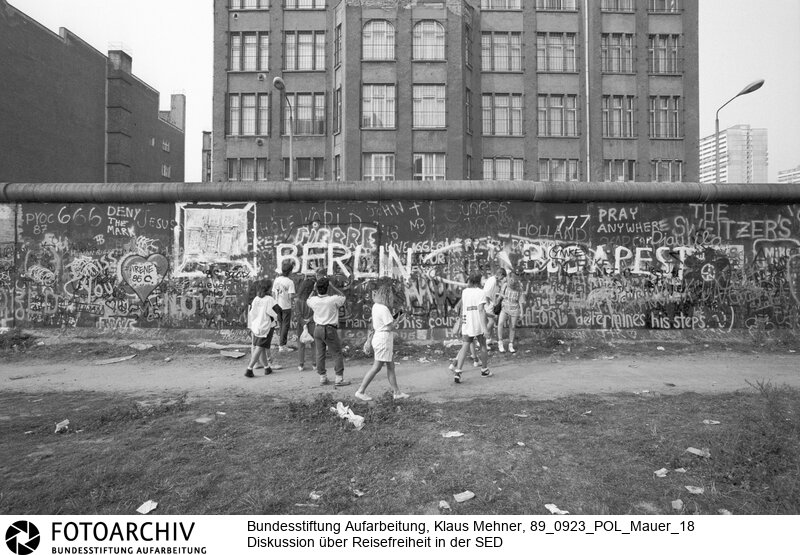 Berlin (Berlin West), 23-09-1989. Diskussion um Reisefreiheit. Über die offene Grenze Ungarns verließen überwiegend junge Familien und Angehörige der technischen Intelligenz die Deutsche Demokratische Republik (DDR). Unter hochrangigen Funktionären der SED (Sozialistischen Einheitspartei) wird der Aderlass und über Reisefreiheit diskutiert. Bilder der Berliner Mauer mit einem Spezialweitwinkel Objektiv. Foto: Jugendliche Touristen an der Mauer, Zimmerstrasse.<BR>Material/Technik: Foto<BR>Berlin (West)<BR>Signatur: 89_0923_POL_Mauer_18