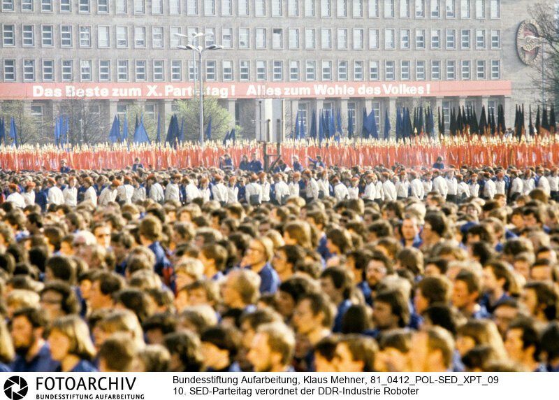 Berlin (Bezirk Berlin) DDR, 12-04-1981. 10. SED-Parteitag verordnet der DDR-Industrie Roboter. Nach Kürzung der Erdöllieferungen durch die Sowjetunion (UdSSR) findet der X. Parteitag der Sozialistischen Einheitspartei Deutschland (SED) (11. bis 16. April) in Anwesenheit des Chefideologen der KPdSU statt. Die Delegierten bestätigten dem Generalsekretär und wählten 156 Mitglieder und 57 Kandidaten für das Zentralkomitee (ZK). In Durchsetzung der Einheit von Wirtschaft- und Sozialpolitik soll der Genosse Industrieroboter in Kombinate und volkseigene Betriebe (VEB) Einzug halten. Foto: Parteinachwuchs Freie Deutsche Jugend (FDJ) und Junge Pioniere (JP) bei Demonstration vor der Parteiführung auf dem Marx-Engels-Platz.<BR>Material/Technik: Foto<BR>Bezirk Berlin<BR>Signatur: 81_0412_POL-SED_XPT_09