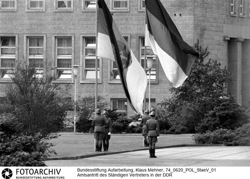 Berlin (Bezirk Berlin) DDR, 20-06-1974. Unterhalb der Anerkennungsschwelle einigten sich die Deutsche Demokratische Republik (DDR) und die Bundesrepublik (BRD) im Grundlagenvertrag auf den Austausch Ständiger Vertreter mit faktisch diplomatischem Status. Foto: Soldaten bei Flaggenparade im Innenhof des Staatsrats.<BR>Material/Technik: Foto<BR>Bezirk Berlin<BR>Signatur: 74_0620_POL_StaeV_01
