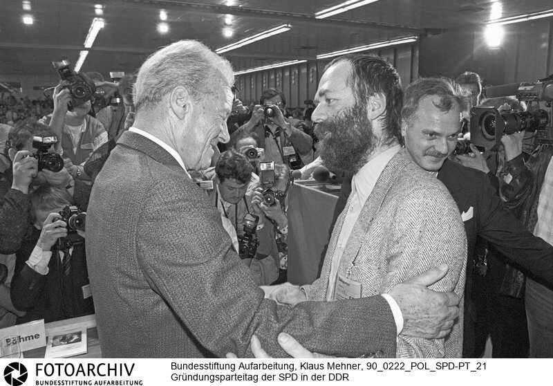 Leipzig (Bezirk Leipzig) DDR, 22-02-1990. Erster Parteitag der SPD in der DDR. Der auf drei Tage angesetzte Parteitag der Sozialdemokratischen Partei (SPD) in der Deutschen Demokratischen Republik (DDR) wählte den Vorstand, verabschiedete das Statut sowie das Programm für die Wahlen zur Volkskammer. Deutschland soll in drei Stufen wieder vereinigt und die Einheit mit einer neuen gesamtdeutschen Verfassung besiegelt werden. Foto: Willy Brandt (77) nimmt die Glückwünsche zum Ehrenvorsitz von Markus Meckel (38) entgegen.<BR>Material/Technik: Foto<BR>Bezirk Leipzig<BR>Signatur: 90_0222_POL_SPD-PT_21