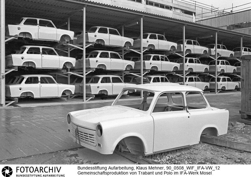 Mosel (Bezirk Chemnitz) DDR, 08. 05. 1990. Gemeinsam ins Chaos. Die Joint venture Produktion der Fahrzeugtypen Viertakttrabant 1.1 von IFA und Polo der Volkswagen AG im Werk der IFA (Industrieverband Fahrzeugbau) startet mit Problemen. Die Autos werden auf dem gleichen Transportsystem gebaut. Außerdem kommt die Polo Vorproduktion nicht voran, da es mit der Zulieferung von Komponenten für den Trabant hapert. Die Projektleitung improvisiert, Polo Karossen werden manuell in das Transportband eingefügt. Foto: Trabant Karosserielager im Freien.<BR>Material/Technik: Foto<BR>Bezirk Chemnitz<BR>Signatur: 90_0508_WIF_IFA-VW_12