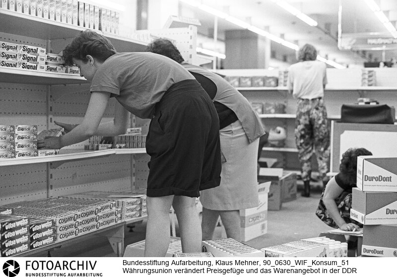 Cottbus (Bezirk Cottbus) DDR, 01. 07. 1990. Volle Regale in der DDR. Die Einführung der D-Mark wird das Preisgefüge und Warenangebot im Osten radikal verändern. Bereits vor der Währungsunion war der Einzelhandel in der Deutschen Demokratischen Republik (DDR) in der Hand westdeutscher Handelskonzerne. Artikel aus heimischer Produktion sind im Warenhaus Konsument mit angegliederter Kaufhalle kaum noch zu finden. Foto: Mitarbeiterinnen bei Auspreisung und Einräumen der Waren.<BR>Material/Technik: Foto<BR>Bezirk Cottbus<BR>Signatur: 90_0630_WIF_Konsum_51