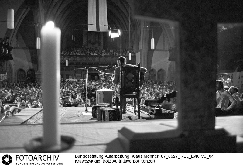 Berlin (Bezirk Berlin) DDR, 27-06-1987. Friedensgruppen und Mitglieder der Kirche von Unten (KvU) halten der Amtskirche zu lasche Interventionen gegen Auflagen des Staatssekretariats für Kirchenfragen der Deutschen Demokratischen Republik (DDR) vor und organisieren ihren eigenen Kirchentag. Die Kirchenleitung sucht mit der Opposition den Konsens und toleriert das Konzert des mit Auftrittsverbot belegten Liedermachers. Foto: Stephan Krawczyk (32) bei Konzert in der überfüllten Samariterkirche.<BR>Material/Technik: Foto<BR>Bezirk Berlin<BR>Signatur: 87_0627_REL_EvKTvU_04