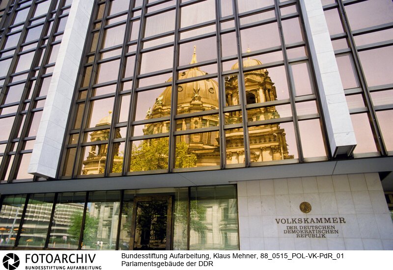 Berlin (Bezirk Berlin) DDR, 15-05-1988. Sitz der DDR-Volkskammer. Das Parlament der Deutschen Demokratischen Republik (DDR) residiert im Palast der Republik (PdR). Der nördliche Teil von Erichs Lampenladen (Volksmund) ist den Abgeordneten der Volkskammer vorbehalten. Foto: Der liebe Gott sieht Alles; in der Thermoverglasung spiegelt sich der Berliner Dom.<BR>Material/Technik: Foto<BR>Bezirk Berlin<BR>Signatur: 88_0515_POL-VK-PdR_01