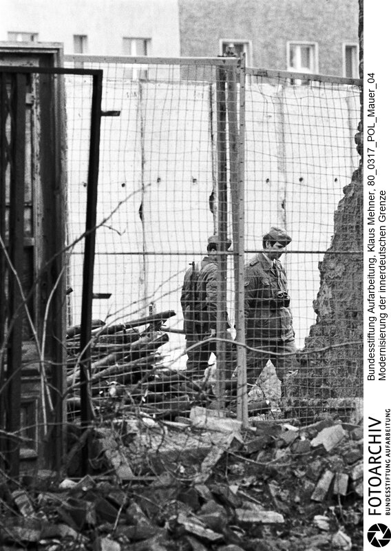Berlin (Berlin West), 17. 03. 1980. Modernisierung der innerdeutschen Grenze. Die Deutsche Demokratische Republik (DDR) baut die Berliner Mauer aus. Die Staatsgrenze West, im Jargon des Regimes auch antifaschistischer Schutzwall genannt, soll höher und noch unüberwindbarer werden. Foto: Soldaten der Grenztruppe bei Patrouille vor den Betonelementen.<BR>Material/Technik: Foto<BR>Berlin (West)<BR>Signatur: 80_0317_POL_Mauer_04