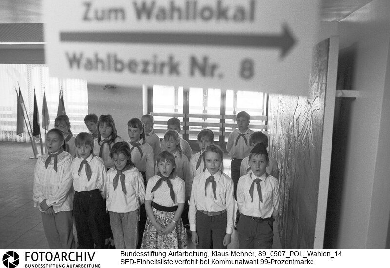 Berlin (Bezirk Berlin) / DDR, 07-05-1989. Die Einheitsliste der Nationalen Front unter Führung der Sozialistischen Einheitspartei Deutschland (SED) fuhr zu den Kommunalwahlen laut offiziellem Endergebnis 98,85 Prozent der Stimmen ein. Beobachter der Opposition und die akkreditierten Journalisten bezweifeln das Ergebnis. Im einzigen Westjournalisten zur Berichterstattung zugewiesenen Wahllokal war die Anzahl der Wahlberechtigten nicht zu erfahren. Foto: Junge Pioniere (JP) erfreuen die Wähler mit Gesang.<BR>Material/Technik: Foto<BR>Bezirk Berlin<BR>Signatur: 89_0507_POL_Wahlen_14