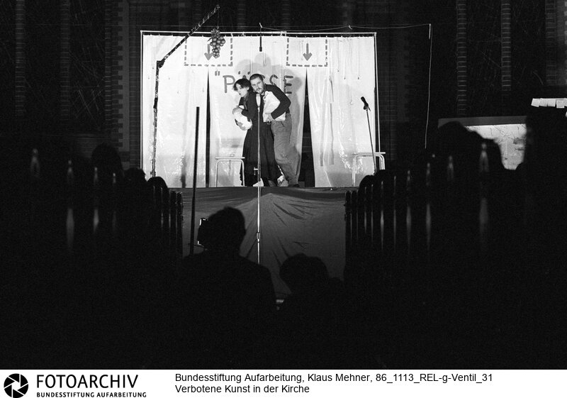 Berlin (Bezirk Berlin) DDR, 13-11-1986. Zum Auftakt der siebenten Friedensdekade verlieh der Pfarrer der evangelischen Samariter Gemeinde das Goldene Ventil an Aktivisten der Friedensbewegung. Anschließend boten im Beisein zahlreicher Mitarbeiter des Ministeriums für Staatssicherheit (MfS, Stasi) mit Auftrittsverbot belegte Künstler einen Farcenabend. Foto: Regisseurin Freya Klier (36), Liedermacher Stephan Krawczyk (31) bei Darbietung ihres Stücks Pässe und Parolen. Copyright by: Klaus Mehner (Higher resolution available)<BR>Material/Technik: Foto<BR>Bezirk Berlin<BR>Signatur: 86_1113_REL-g-Ventil_31