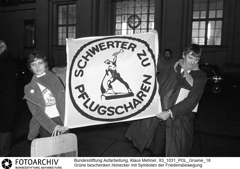 Berlin (Bezirk Berlin) DDR, 31-10-1983. Der Staatsratsvorsitzende der Deutschen Demokratischen Republik (DDR) empfing Fraktions- und Vorstandsmitglieder der Grünen. Die Delegation der Igelpartei bedachte den Chef der Sozialistischen Einheitspartei Deutschland (SED) mit einem Friedensvertrag und dem Foto der Skulptur Schwerter zu Pflugscharen. Anschließend demonstrierten die Aktivisten mit in der DDR verbotenen Symbolen der Friedensbewegung vor dem Staatsrat. Foto: Petra Kelly, Otto Schily (Die Grünen) bei Demonstration vor dem Staatsrat.<BR>Material/Technik: Foto<BR>Bezirk Berlin<BR>Signatur: 83_1031_POL_Gruene_18