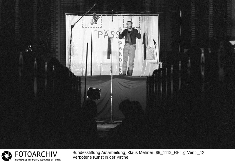 Berlin (Bezirk Berlin) DDR, 13-11-1986. Zum Auftakt der siebenten Friedensdekade verlieh der Pfarrer der evangelischen Samariter Gemeinde das Goldene Ventil an Aktivisten der Friedensbewegung. Anschließend boten im Beisein zahlreicher Mitarbeiter des Ministeriums für Staatssicherheit (MfS, Stasi) mit Auftrittsverbot belegte Künstler einen Farcenabend. Foto: Liedermacher Stephan Krawczyk (31) bei Darbietung des Stücks Pässe und Parolen. Copyright by: Klaus Mehner (Higher resolution available)<BR>Material/Technik: Foto<BR>Bezirk Berlin<BR>Signatur: 86_1113_REL-g-Ventil_12