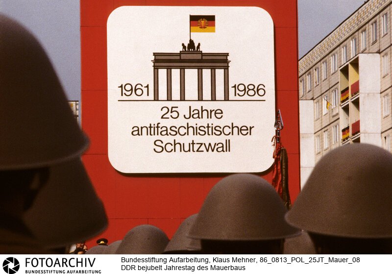 Berlin (Bezirk Berlin) DDR, 13-08-1986. Mit einem Kampfappell der Kampfgruppen (KG) der Arbeiterklasse beging die Deutsche Demokratische Republik (DDR) den 25. Jahrestag des Baus des Antifaschistischen Schutzwalls, vulgo Mauer. Der SED-Chef würdigte das Bauwerk als Voraussetzung für die Friedens- und Entspannungspolitik. Alsdann nahmen Partei- und Staatsführung den Vorbeimarsch von Soldaten der Nationalen Volksarmee (NVA) und Betriebskampfgruppen ab. Foto: Festtagssignet der Tribüne.<BR>Material/Technik: Foto<BR>Bezirk Berlin<BR>Signatur: 86_0813_POL_25JT_Mauer_08